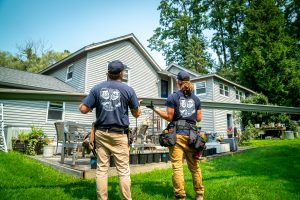 Two The Brothers That Just Do Gutters gutter installers hold a long gutter in front of a white house they are about to install the gutter on.